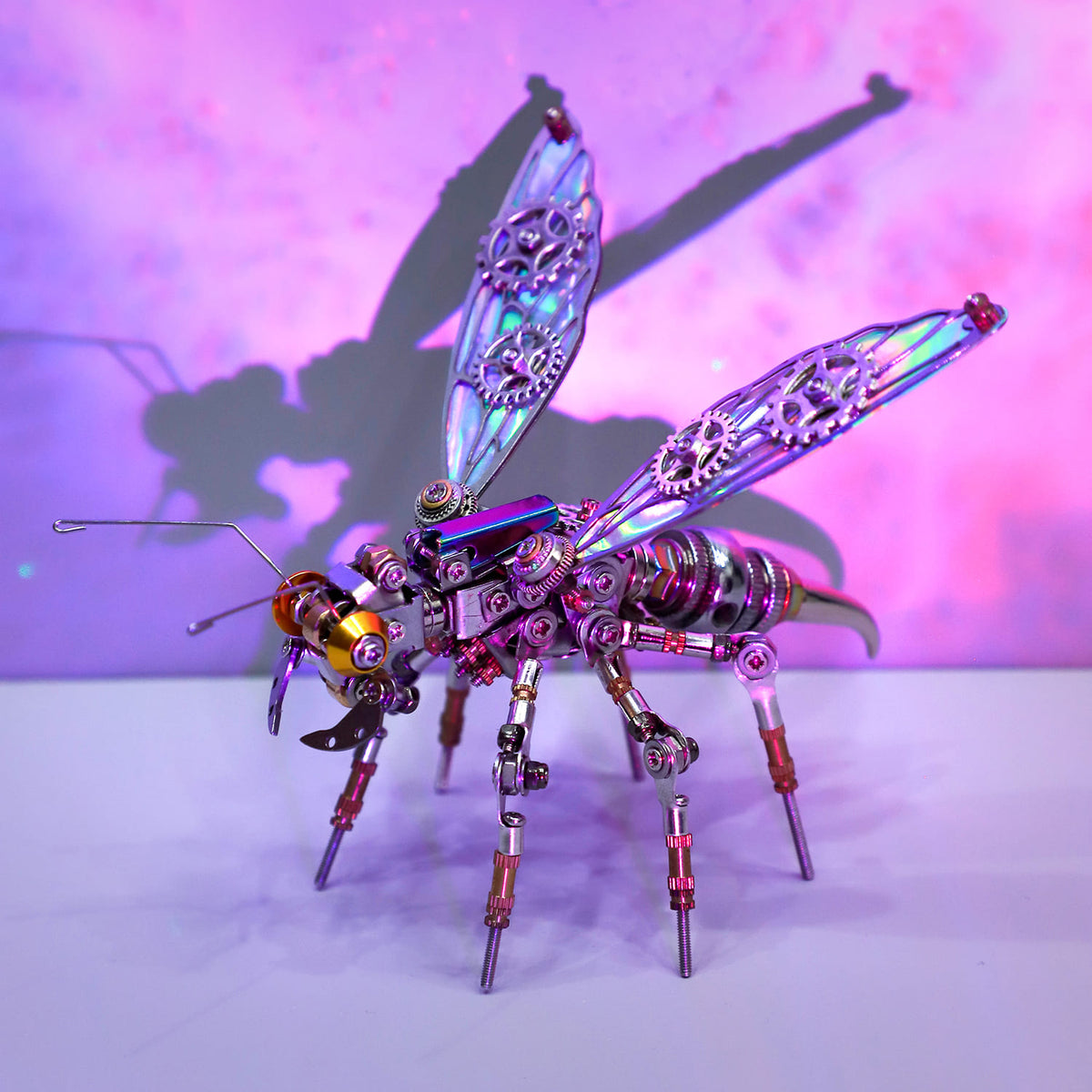 Metal insect sculpture with iridescent wings on a white surface with a purple gradient background
