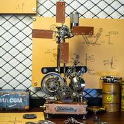 A steampunk-style mechanical sculpture or model with gears, metal parts, wooden panels, and a small engine-like assembly on a tabletop. The setup is photographed in a workshop-like environment with a chain-link wall, yellow schematic boards, and industrial containers in the background.