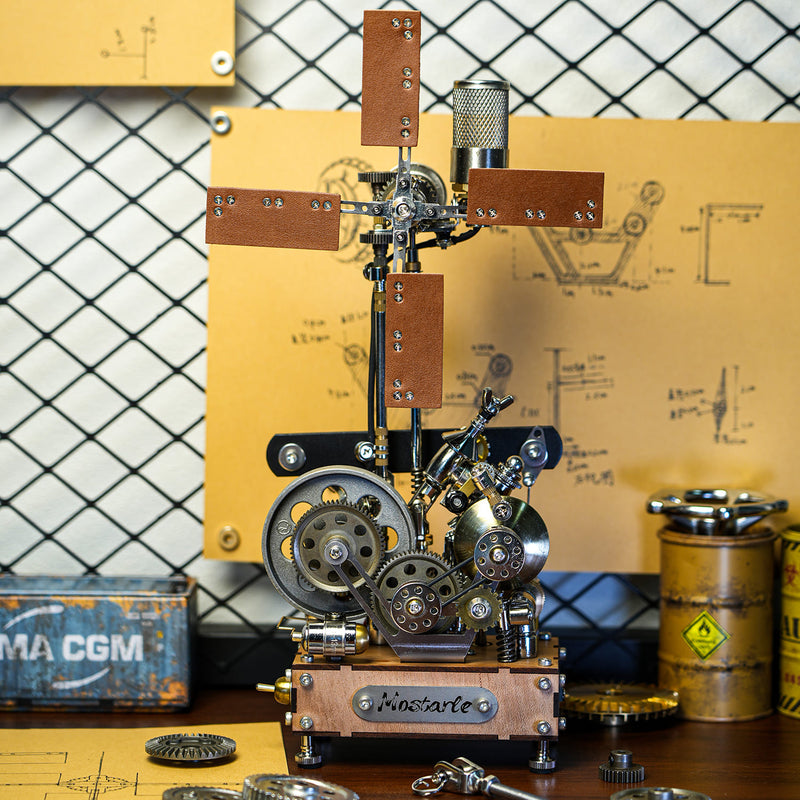 A steampunk-style mechanical sculpture or model with gears, metal parts, wooden panels, and a small engine-like assembly on a tabletop. The setup is photographed in a workshop-like environment with a chain-link wall, yellow schematic boards, and industrial containers in the background.