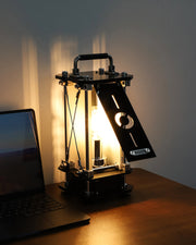 Vintage-style lamp with a glowing light bulb on a wooden surface next to a laptop.