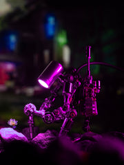 A mechanical model with a pinkish hue, featuring intricate details and a small lamp illuminating part of it, set against a blurred background with a plant.