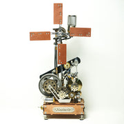 A steampunk-style mechanical model on a white background, featuring gears, cogs, a windmill-like cross, brass-toned metal parts, and a wooden base labeled 'Miostrale'.