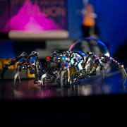 Metallic spider sculpture with colorful details on a reflective surface, blurred background