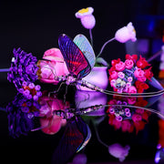 Colorful butterfly-shaped object with a reflective surface, surrounded by flowers on a dark background