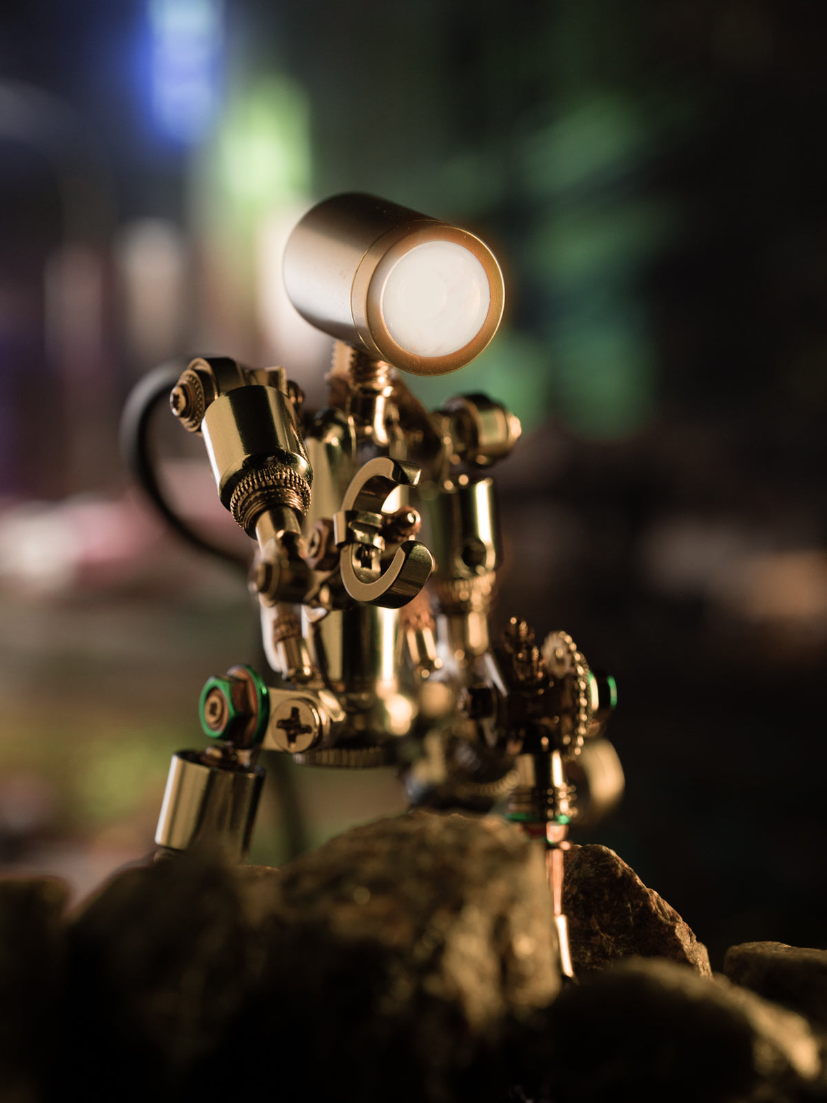A mechanical model with a pinkish hue, featuring intricate details and a small lamp illuminating part of it, set against a blurred background with a plant.