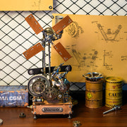 A steampunk-style mechanical model with gears, metal parts, and two wooden blades, displayed on a tabletop against a yellow schematic backdrop with industrial props like barrels and a sign.