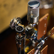 Close-up of a steampunk-style mechanical device with silver metal gears, a mesh-covered cylindrical component, and brown wooden plates against a warm blurred background.